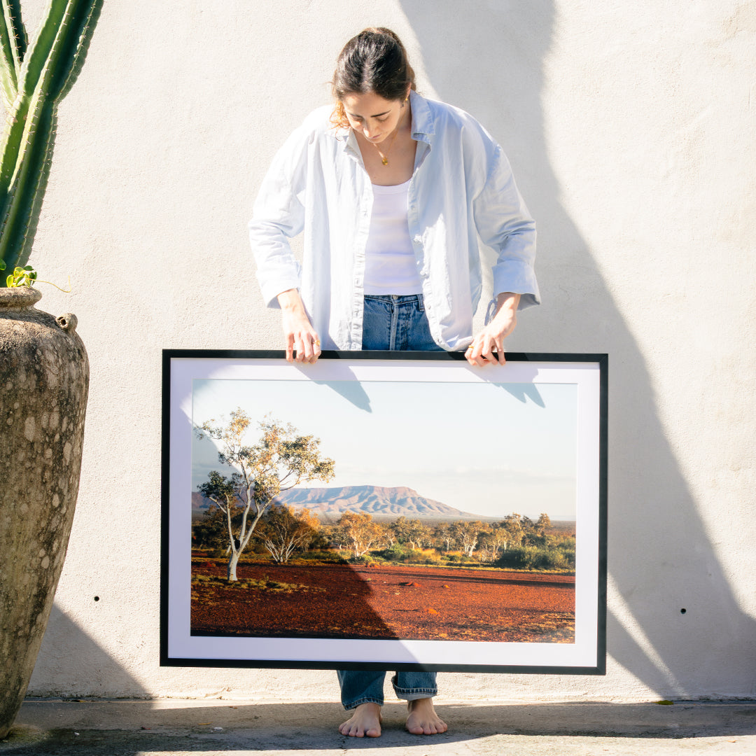 female holding a black photo frame with mountain, tree and red soil